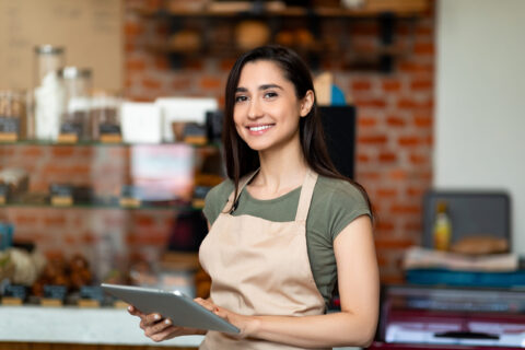 Opening small business. Happy arab woman in apron near bar counter holding digital tablet and looking at camera